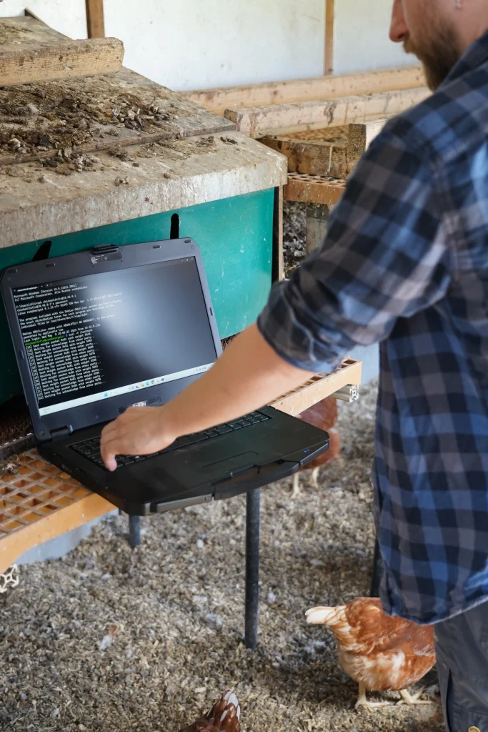 Entwickler steht im Hühnerwagen an einem Computer. Im Hintergrund sind die Hühner zu sehen.